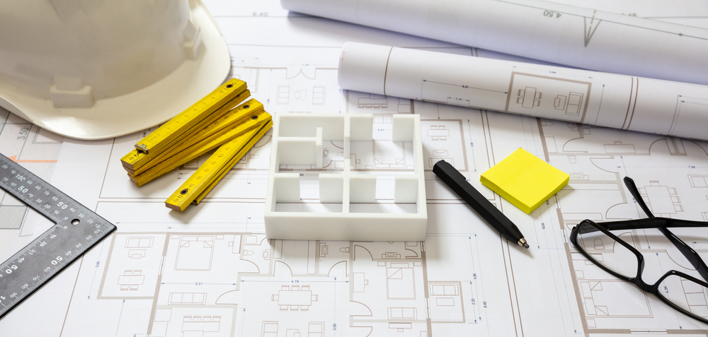 Image of project blueprints with items on top, including a pen, hard hat, and house model, representing the design and build process.