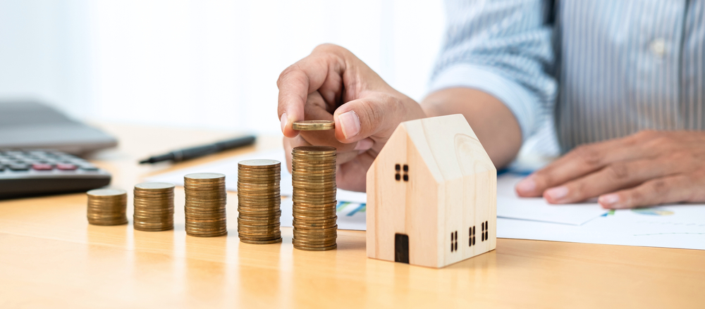 Image of a model wooden house next to piles of coins, representing planning and budgeting for a custom build home.