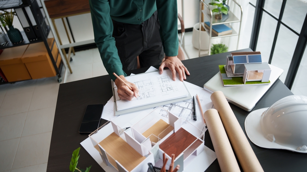Image of a person drawing on architectural plans on a table, next to a model house.